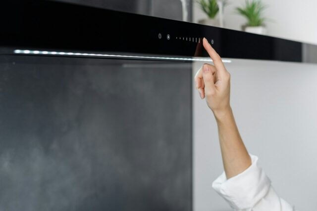 Hand reaching up to press the touch controls on a modern black cooker hood mounted above a hob, with soft lighting and small plants in the background.